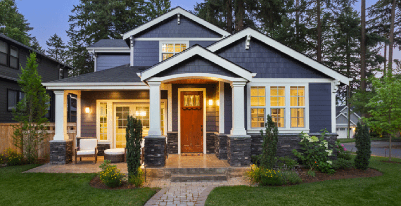 Blue and white two story home at night with house lights on, a green lawn in front of the home and a forest behind the home.